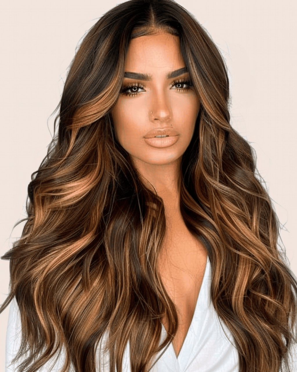 A woman with a full, voluminous hair style showcasing a lace wig named Isabella. The wig has a rich, dark brown root that gradually transitions into a blend of caramel and honey blonde highlights towards the mid-lengths and ends. The hair is styled in soft, flowing waves that create a luxurious and dynamic look, with face-framing layers that start at the chin and add structure to the hairstyle.